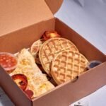 Cardboard takeout box featuring waffles, omelette, and grilled tomatoes, perfect breakfast meal.